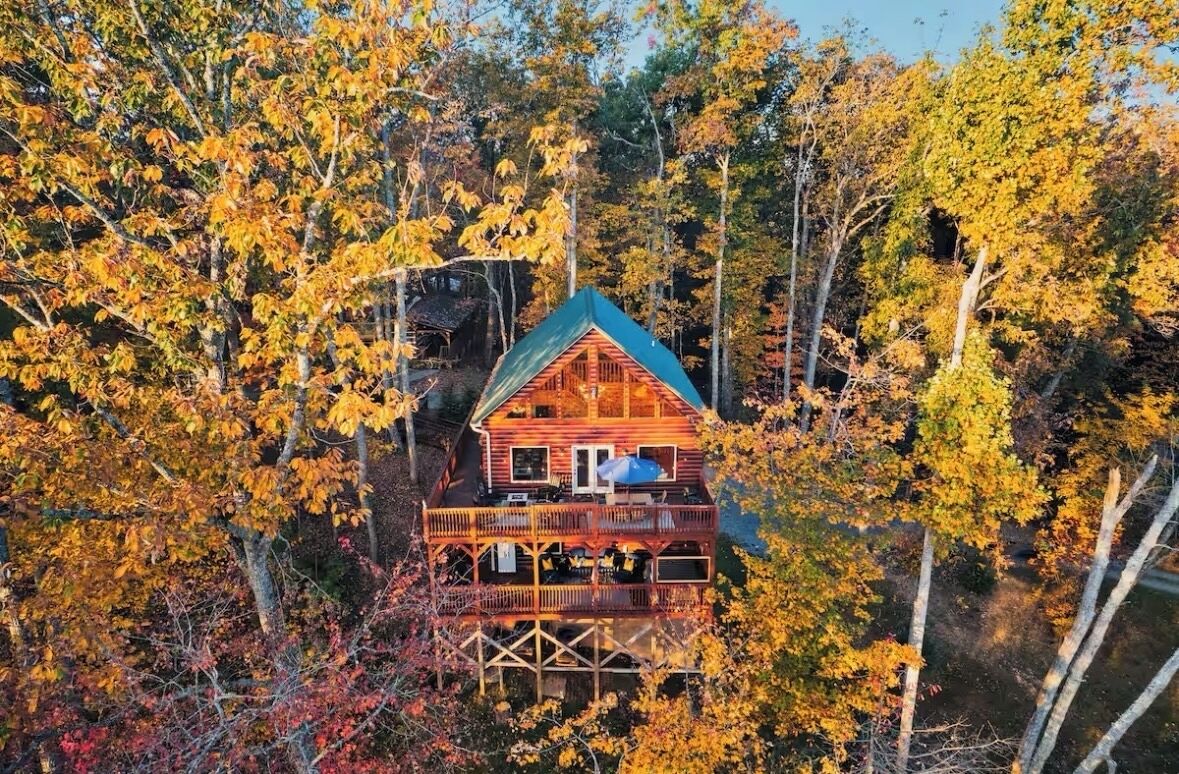 Eine Hütte, umgeben von Herbstlaub, mit einer großen Terrasse und einem steilen Satteldach.