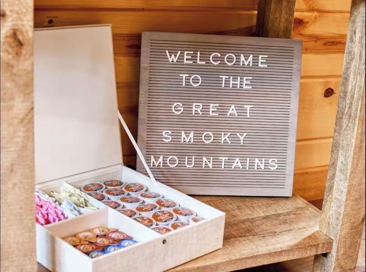 Ein Holzblockhaus-Interieur mit einem Schild, auf dem 'WELCOME TO THE GREAT SMOKY MOUNTAINS' steht, und einer Kiste mit verschiedenen Snacks.
