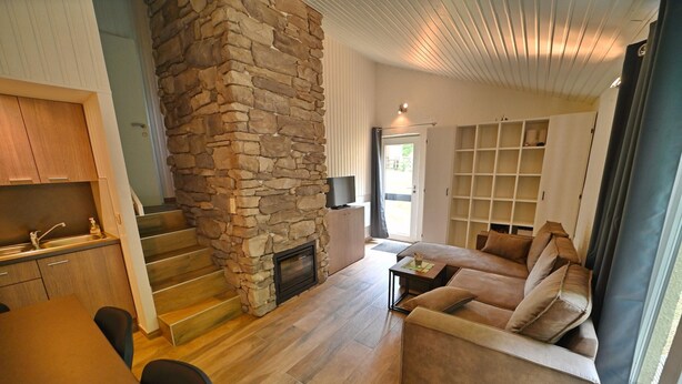 A modern living room with a stone fireplace, a sofa, and a dining area with a kitchen in the background. - Bungalow in Virton by Vallée de Rabais Lake (VIRTON)