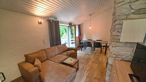A modern living room with a brown leather sofa, a wooden coffee table, a stone accent wall, and a dining area with a table and chairs. - Bungalow in Virton by Vallée de Rabais Lake (VIRTON)