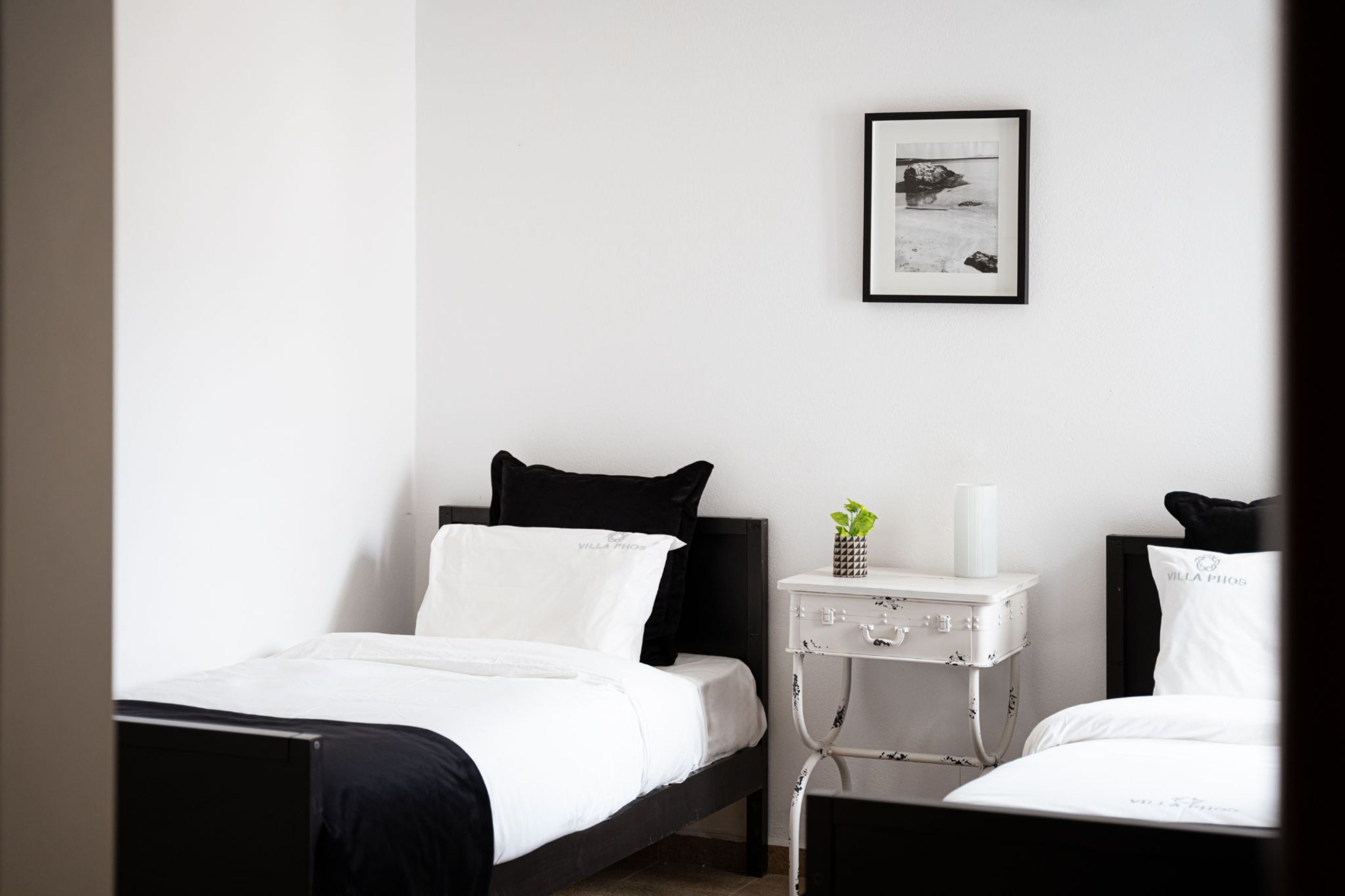 Une chambre d’hôtel au style minimaliste, avec deux lits, du linge de lit blanc, des têtes de lit noires, une table de chevet agrémentée d’une plante et un tableau encadré au mur.