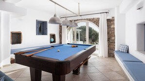 A pool table in a room with a stone wall, a bench, and a view of the outdoors. - Villa Phos Paros (Paros)