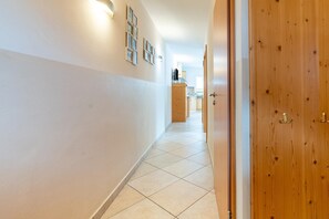 A hallway with a wooden door on the right, a tiled floor, and a kitchen area in the distance.