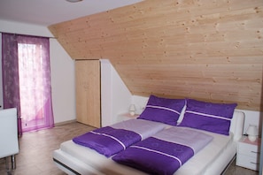 A bedroom with a wooden ceiling, a bed with purple bedding, a white bedside table, and a small nightstand.