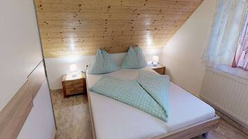 Ein Schlafzimmer mit schrägen Wänden, einem hölzernen Kopfteil, einem Bett mit gemusterten Betttextilien, Nachttischlampen und einem Fenster mit Vorhängen.
