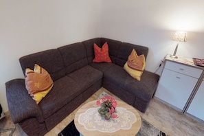 A corner living room with a dark brown sofa, two decorative pillows, a small round coffee table with a floral centerpiece, and a side table with a lamp.