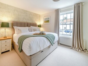 A neatly made bed with pillows, a bedside table with a lamp, a window with curtains, and a wall with a floral pattern.