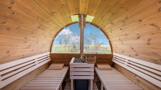 A sauna with wooden benches and a central heating column, surrounded by wooden walls and a large arched window. - Charming Cottage with Sauna (Honnelles)