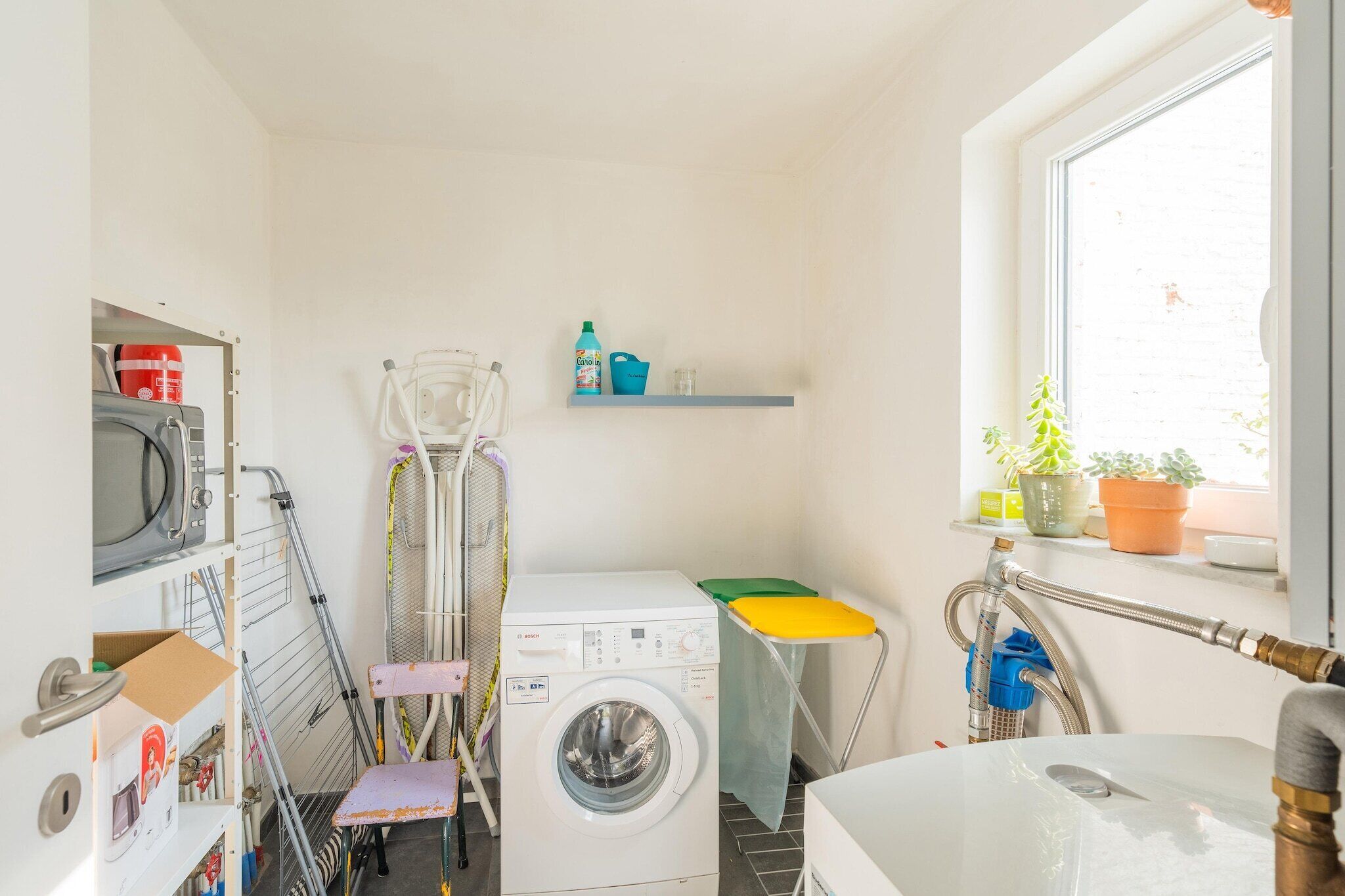 Une petite buanderie comprenant un lave-linge, des étendoirs et une petite étagère où sont rangés les produits d’entretien.