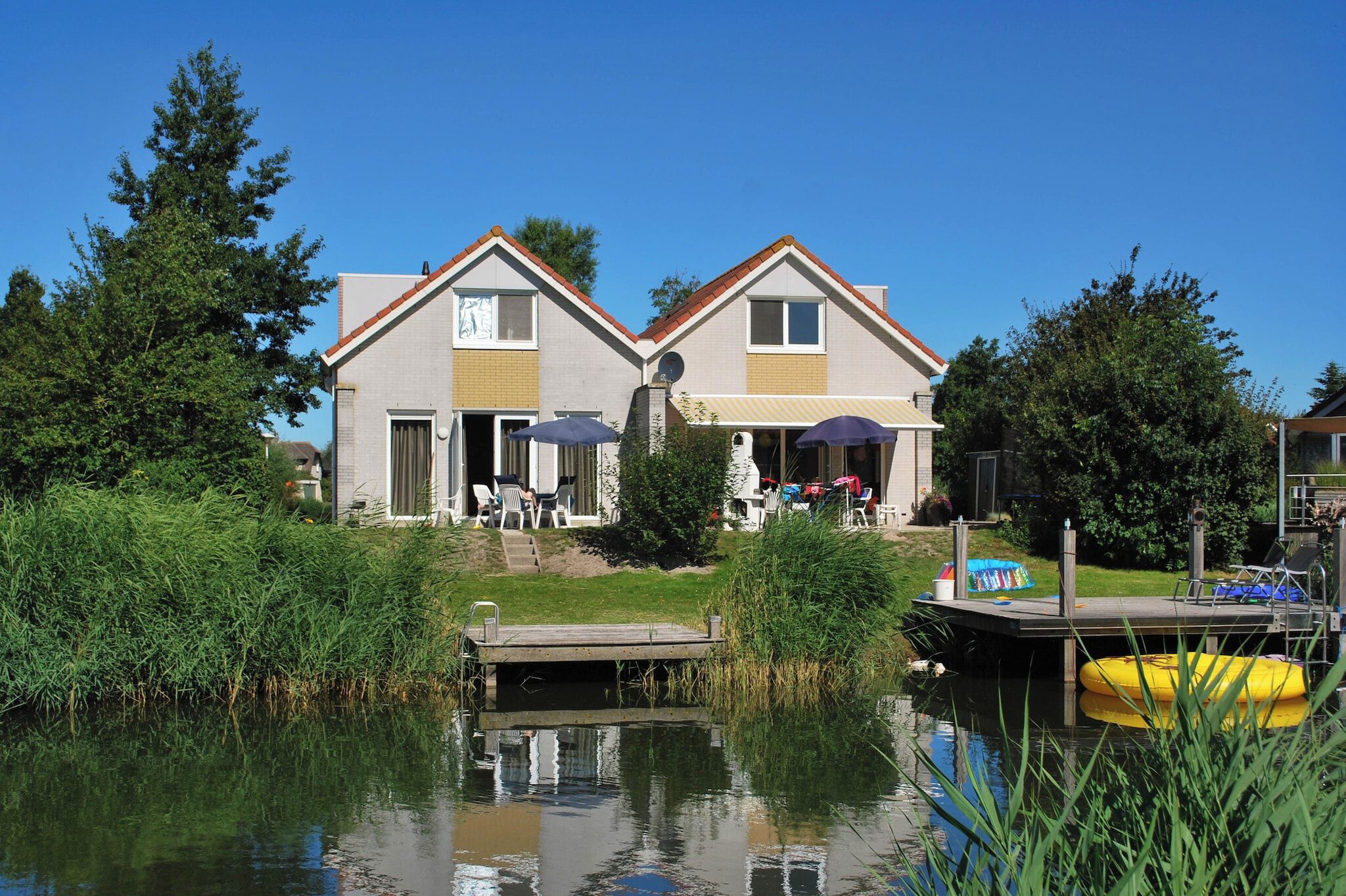 A two-story house with a dock extending into a calm body of water, surrounded by greenery.
