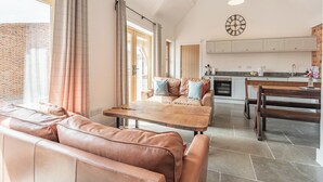 A spacious living area with a large window, a wooden dining table, a leather sofa, and a kitchen in the background. - The Red Rooster - UK44440 (Arlingham, near Frampton-on-Severn)