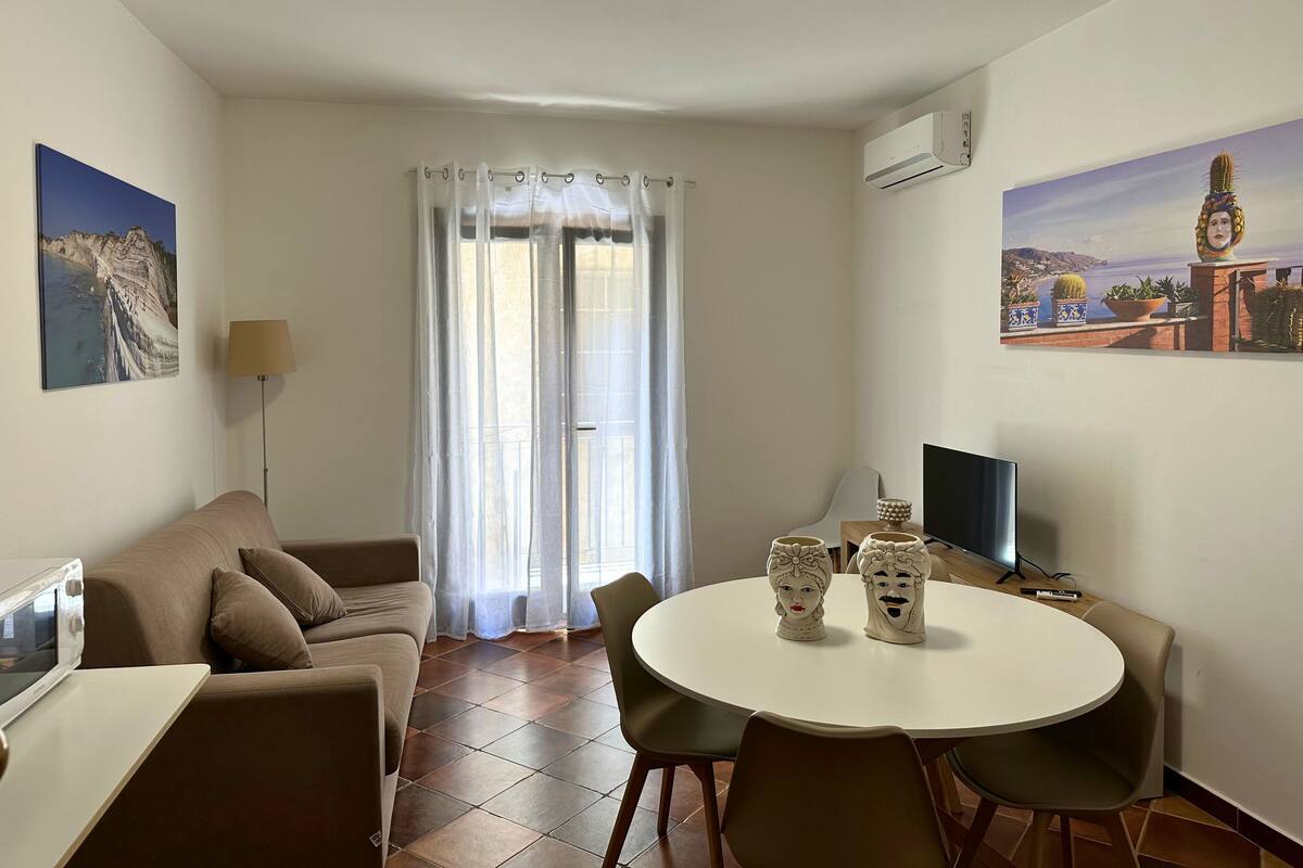 A living room with a sofa, a dining table with chairs, a TV, and a window with curtains.