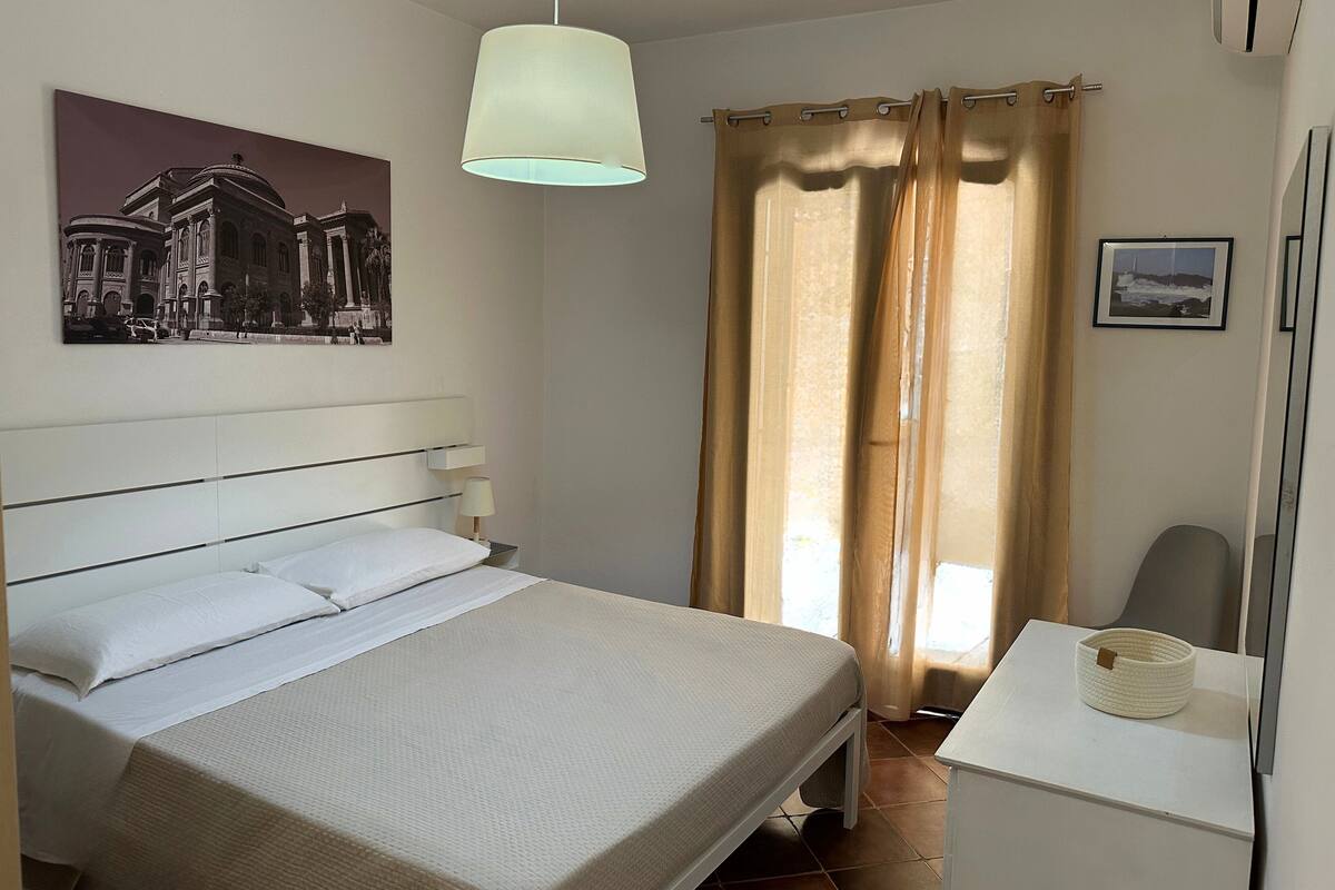 A hotel room with a bed, a desk, a lamp, a picture on the wall, and a window with curtains.