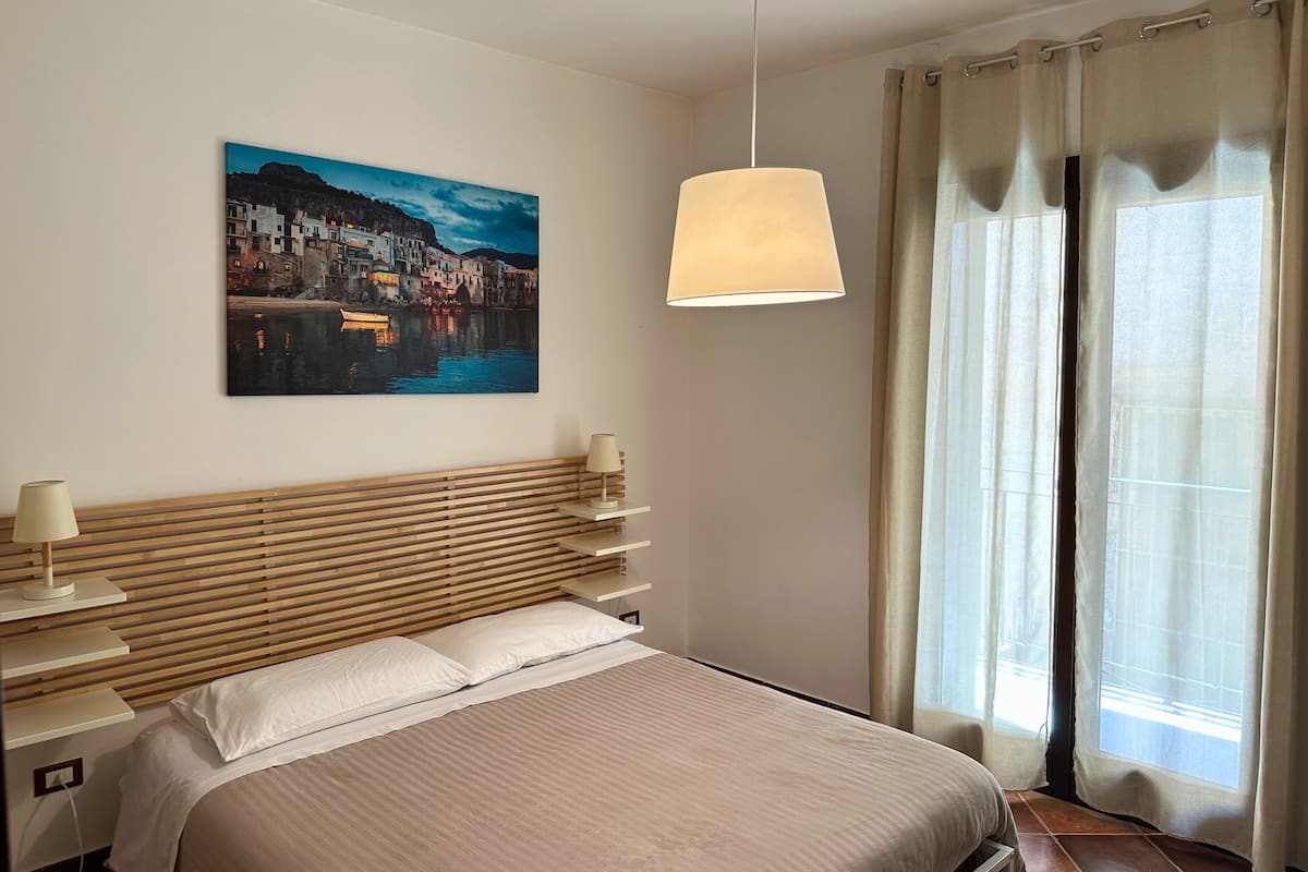 A bedroom with a bed, a nightstand, a lamp, a painting of a coastal town, and a window with curtains.
