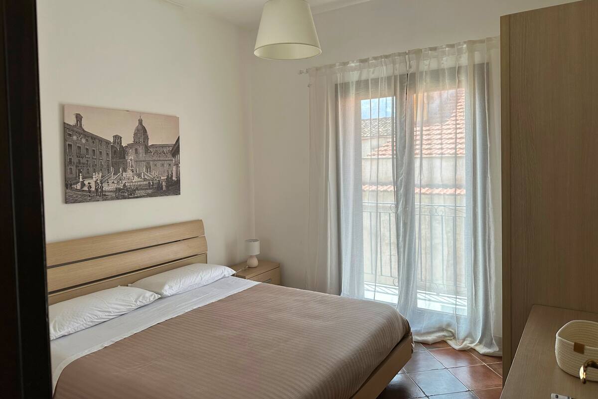 A bedroom with a bed, a nightstand, a lamp, a window with sheer curtains, and a wall-mounted picture.
