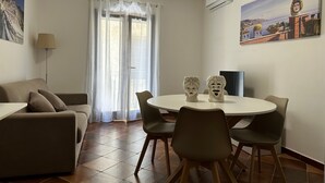 Una sala de estar con un sofá, una mesa de comedor con sillas, una ventana con cortinas, una lámpara y un cuadro en la pared.