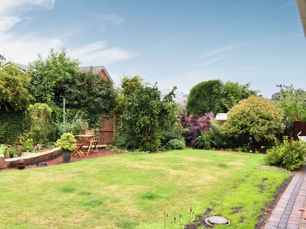 A well-maintained garden with a wooden deck, a variety of plants, and a clear sky.