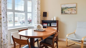Un coin repas avec une table ronde en bois, quatre chaises, un radiateur, une fenêtre avec des rideaux, une bibliothèque et un tableau accroché au mur.