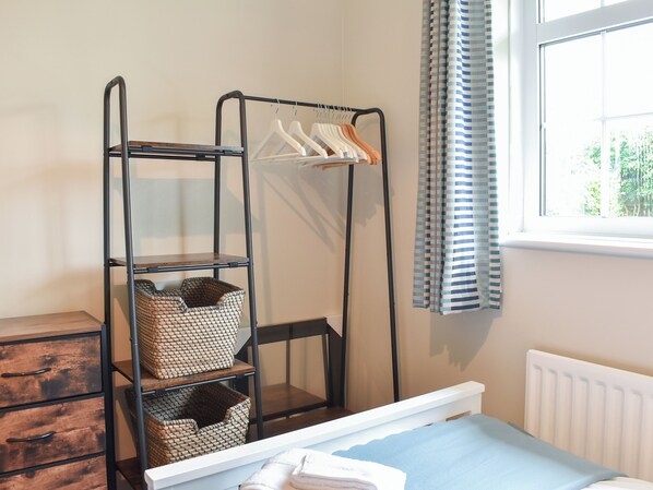A room with a bed, a dresser, a metal shelf unit with baskets, a clothing rack, and a window with curtains.