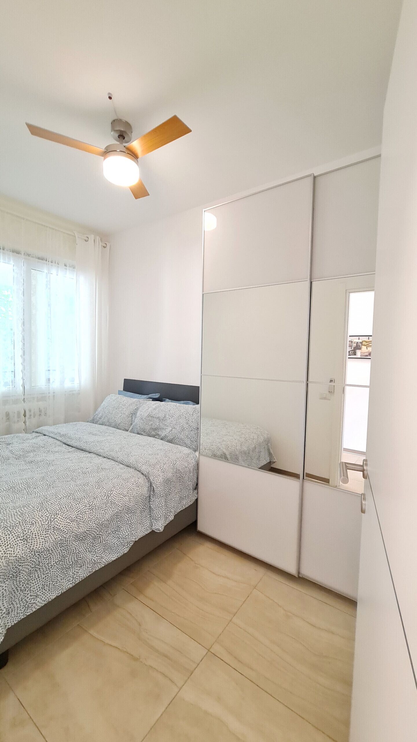 Ein Schlafzimmer mit einem Bett, einem Deckenventilator, einem großen Spiegel und einem Fenster mit Vorhängen.