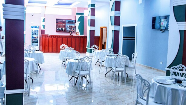 A restaurant with white tables and chairs, a red counter, and a TV on the wall. - Fono Hôtel (Yaoundé)