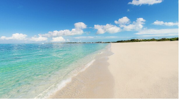 A beach with clear turquoise water, a sandy shore, and a distant shoreline with buildings.