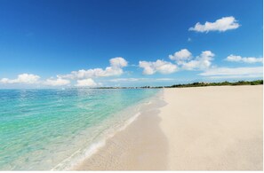 Une plage à l’eau turquoise et limpide, un rivage sablonneux et une ligne côtière lointaine ponctuée de bâtiments.