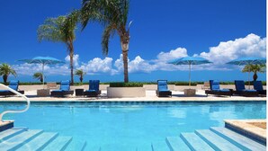 A swimming pool with clear blue water, surrounded by lounge chairs and umbrellas, with palm trees and a clear sky in the background.