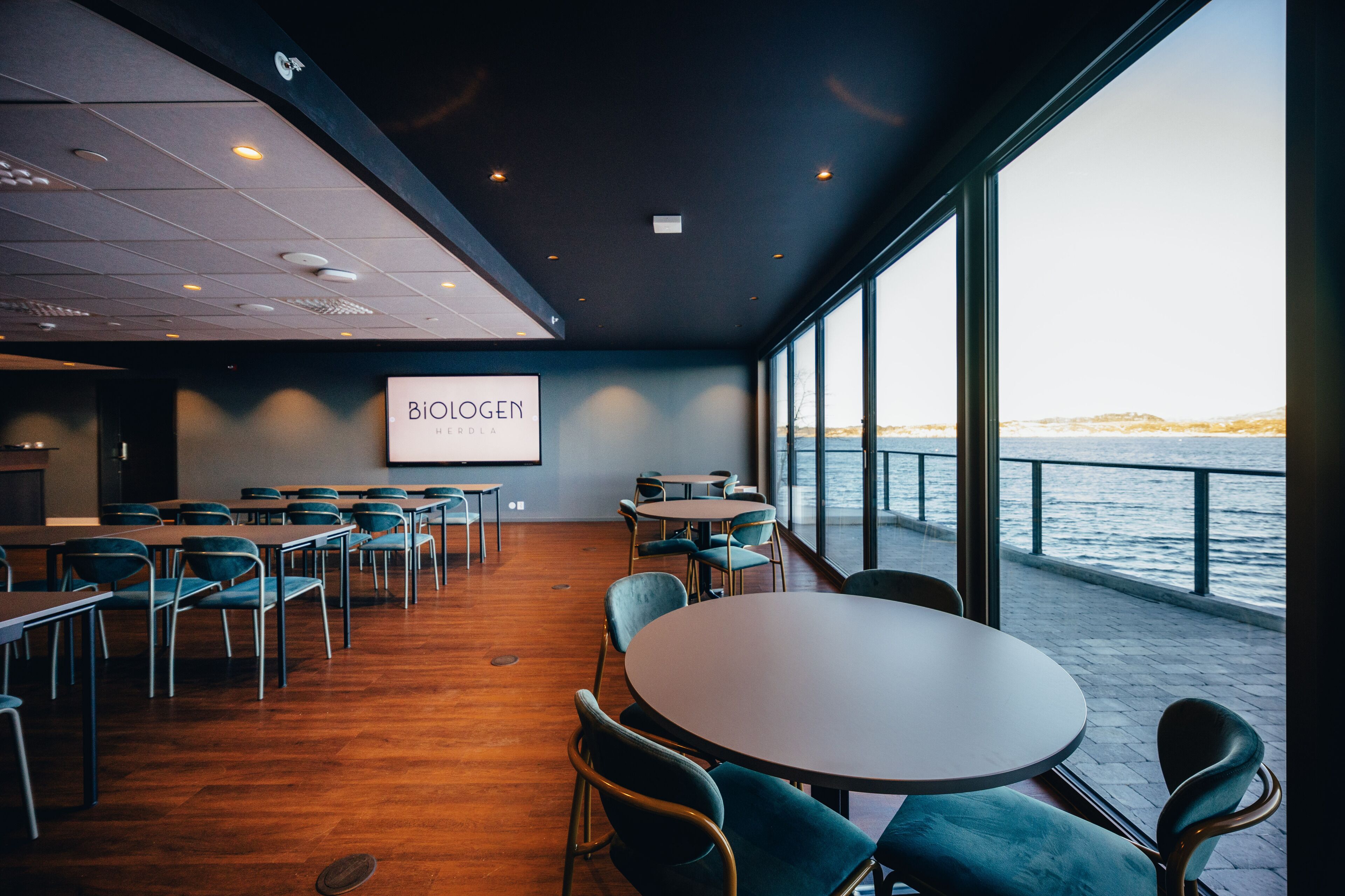 A conference room with a large screen, tables, and chairs, overlooking a waterfront.