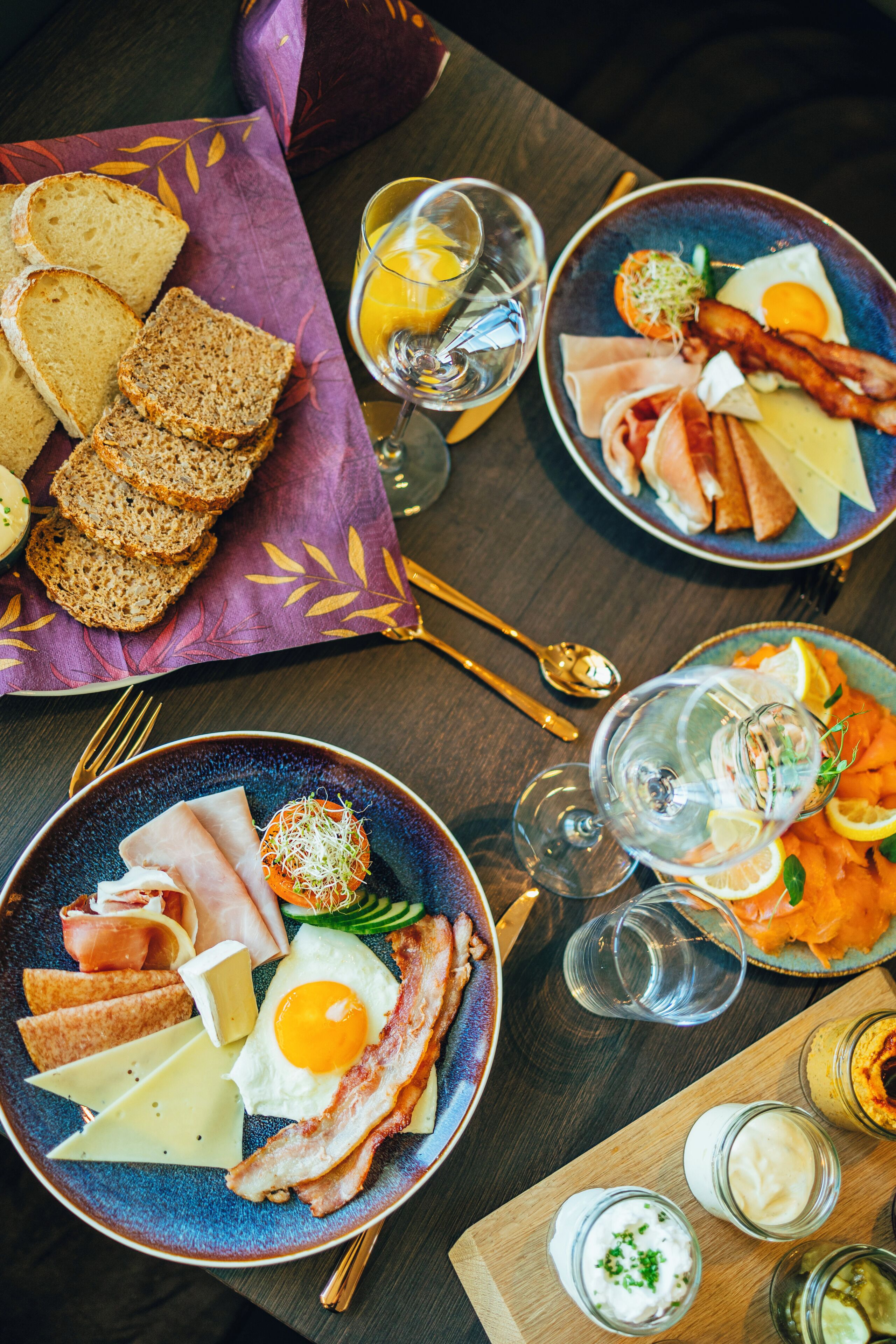 Une table dressée pour le petit-déjeuner avec des assiettes contenant des œufs, du bacon, du jambon, du fromage et des toasts, ainsi que des verres de jus d’orange et d’eau.