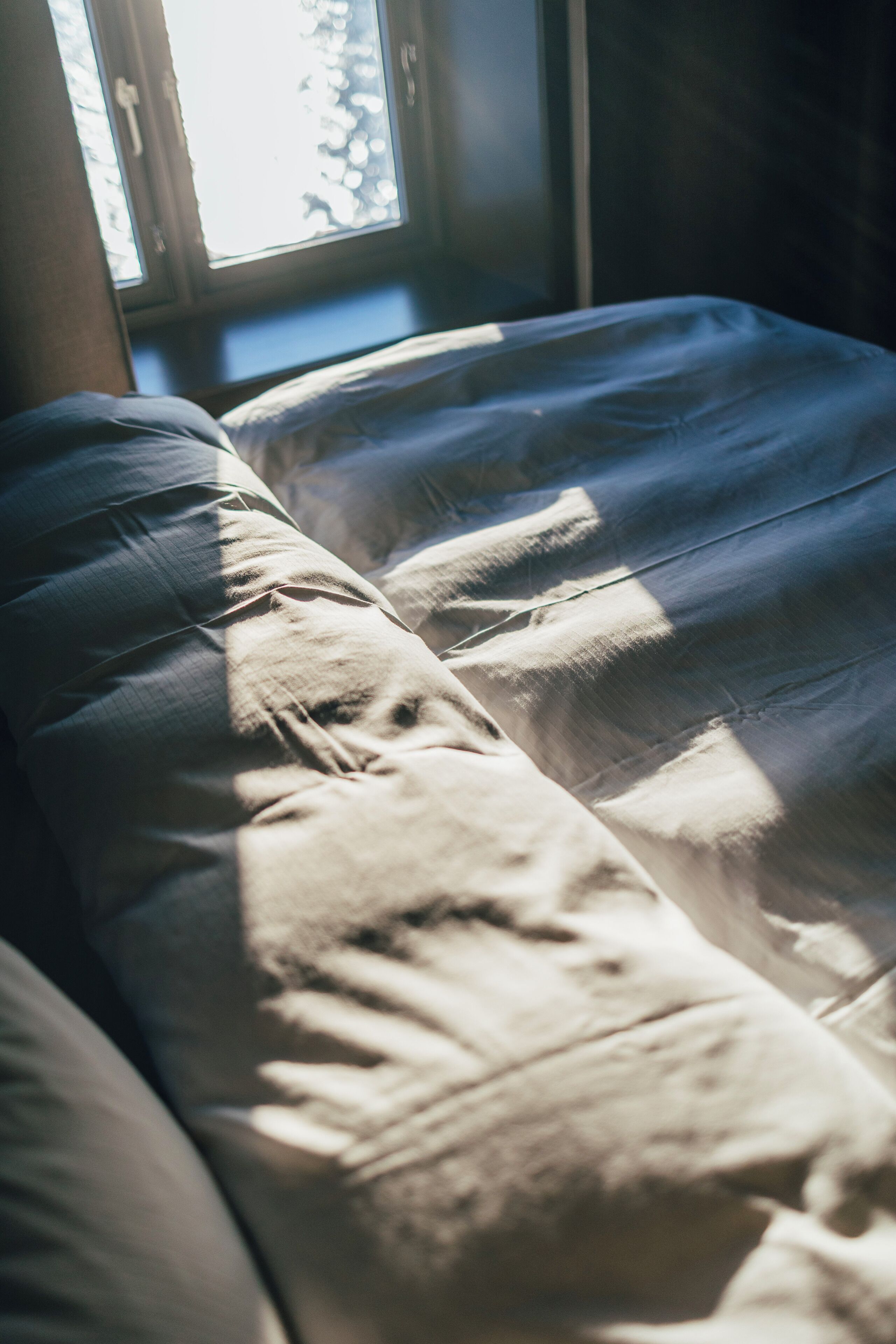 Un lit recouvert d’une couverture blanche, avec un oreiller, placé près d’une fenêtre où la lumière du soleil projette des ombres.