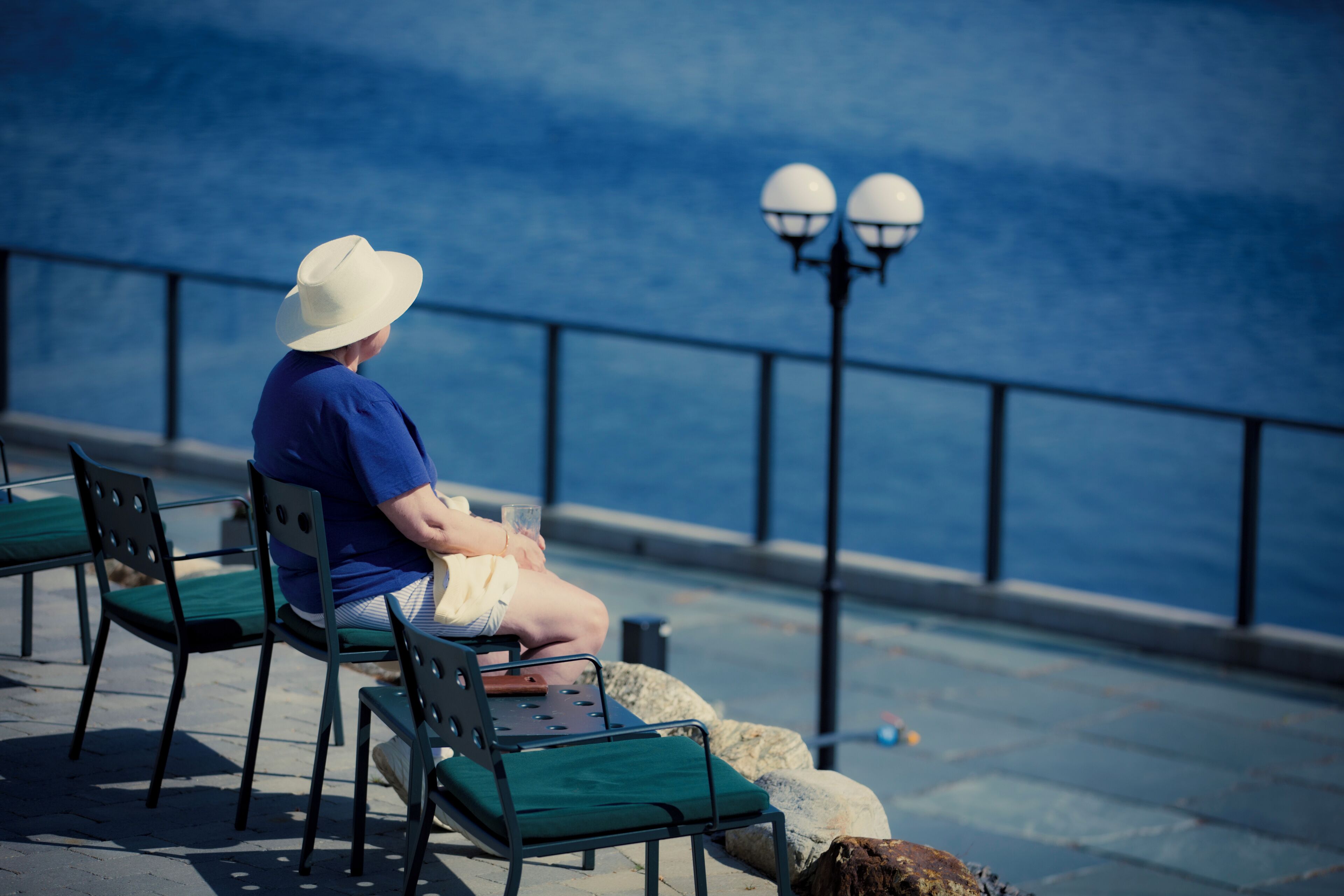 A person sitting on a bench by the water, with a railing and street lamps in the background.