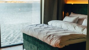 Una cama con ropa blanca y cabecera verde, ubicada junto a una ventana con vista al mar.