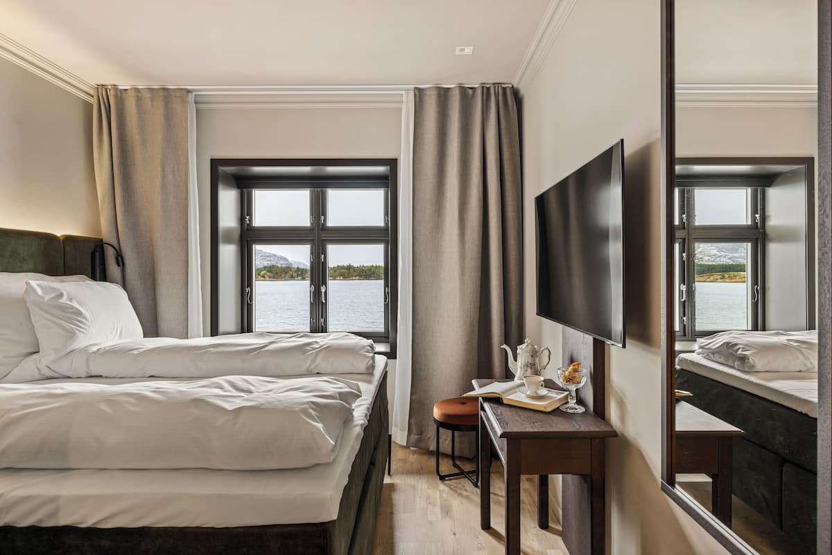 Ett hotellrum med en säng, ett skrivbord med en stol, en tv och ett fönster med utsikt över en vattenyta.