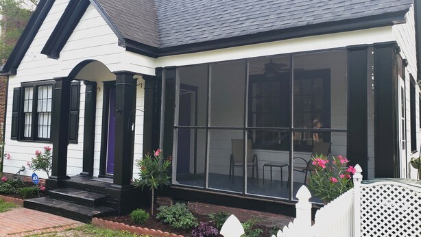 Une maison blanche avec un toit noir, une grande véranda dotée de larges fenêtres et un chemin en briques menant à l’entrée.