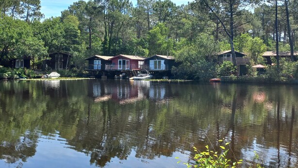A lakeside property with two houses, one with a red roof and another with a brown roof, surrounded by dense trees and calm water reflecting the structures.