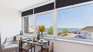 A room with a dining table and chairs, a window with a view of the sea and buildings, and a vase with flowers on the windowsill. - Swn Y Mor - 1 Bedroom Studio - Saundersfoot (Saundersfoot)