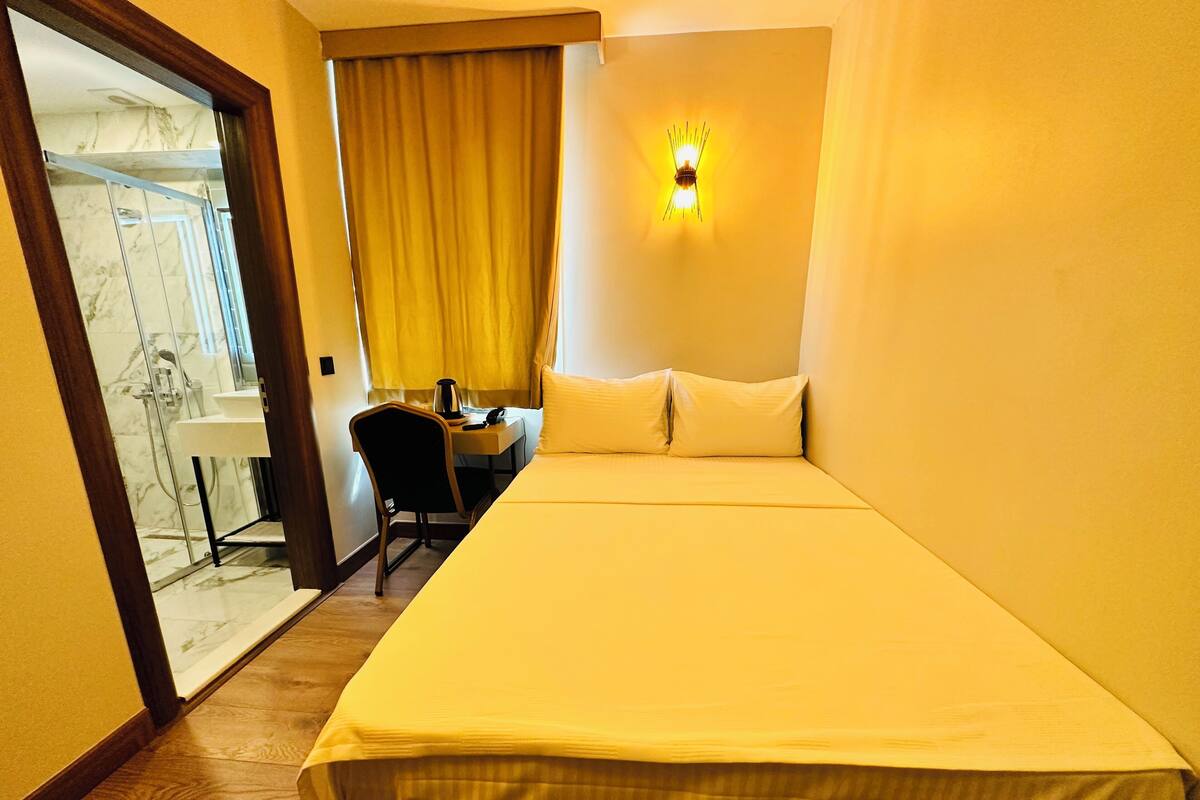 A hotel room with a bed, a desk, a chair, a window with curtains, and a shower area.