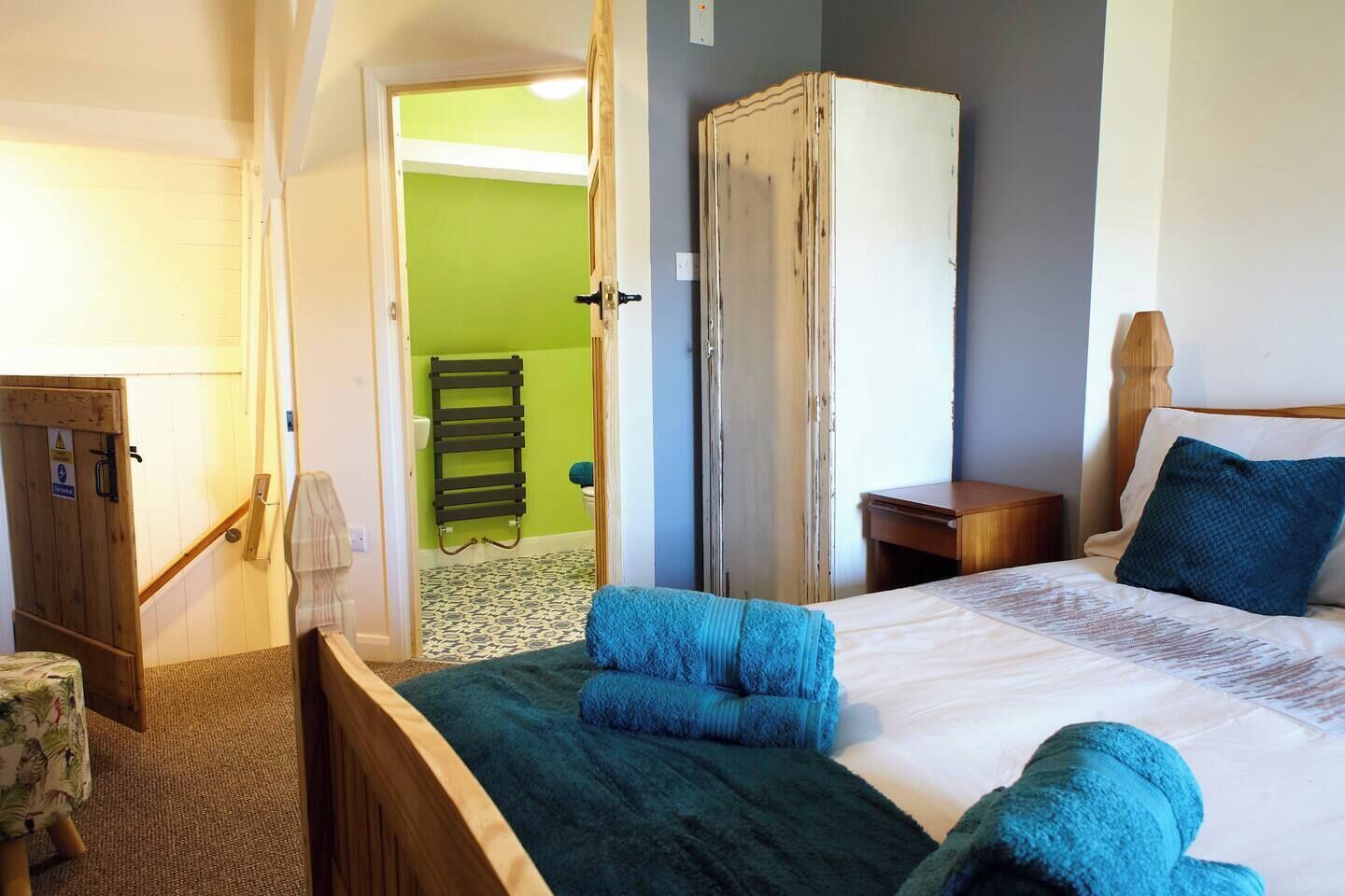 A hotel room with a bed, a wooden wardrobe, a nightstand, a bedside table, and a bathroom visible through an open door.