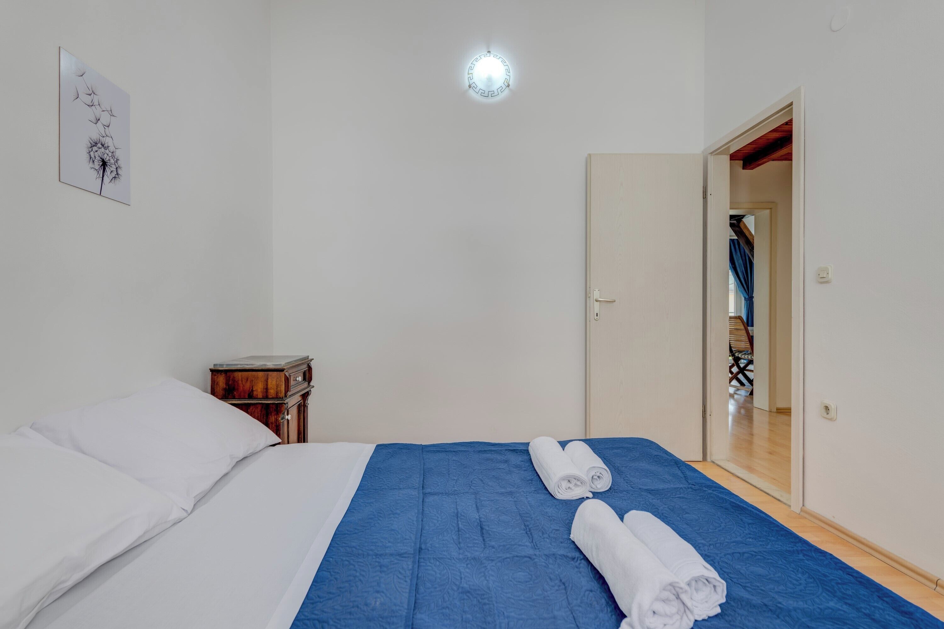 Una cama bien hecha con colcha azul y almohadas blancas, una mesita de noche de madera y una lámpara fijada a la pared.