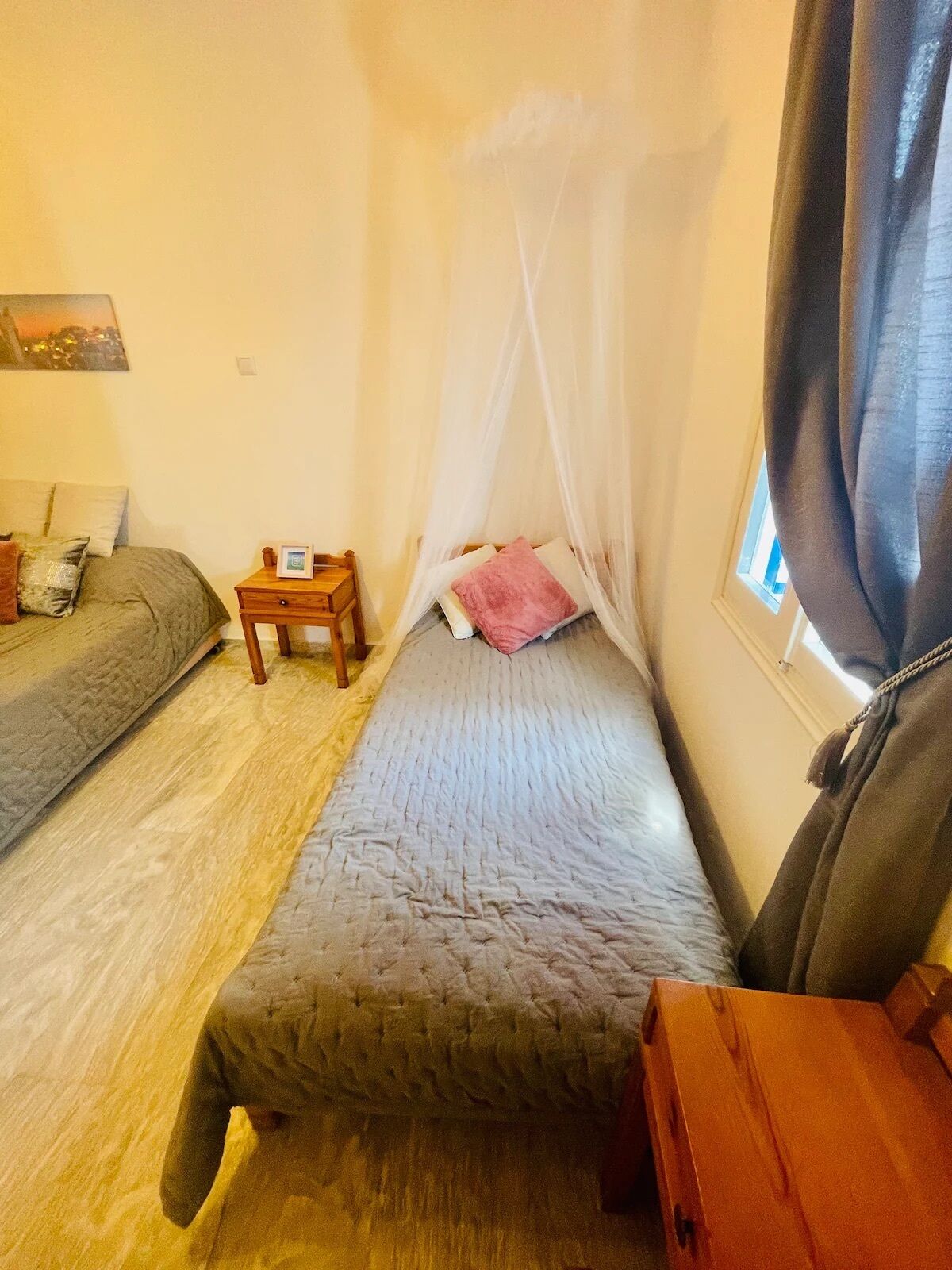 A small, cozy room with two single beds, a bedside table, a nightstand, and a window with curtains.