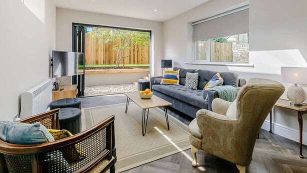 A modern living room with a sofa, armchairs, a coffee table, and a TV. There is a large window with a view of a wooden fence and trees.