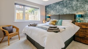 A bedroom with a large bed, a wicker chair, a nightstand with lamps, and a window with a view of a wooden fence.