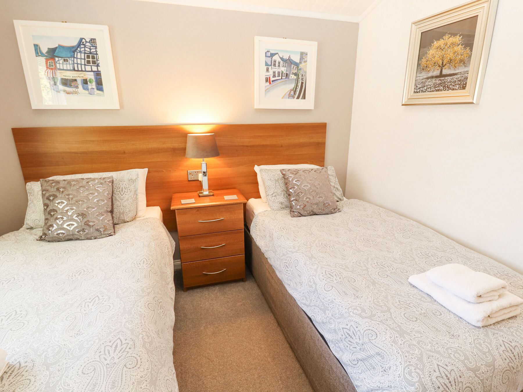 A hotel room with two beds, a wooden headboard, a nightstand with a lamp, and three framed pictures on the walls.