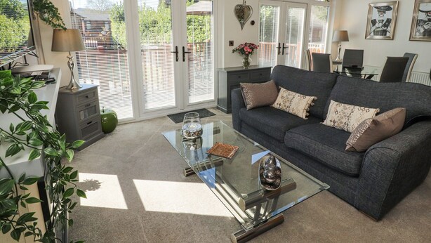 A modern living room with a grey sofa, glass coffee table, and a view through large sliding glass doors to the outdoors.