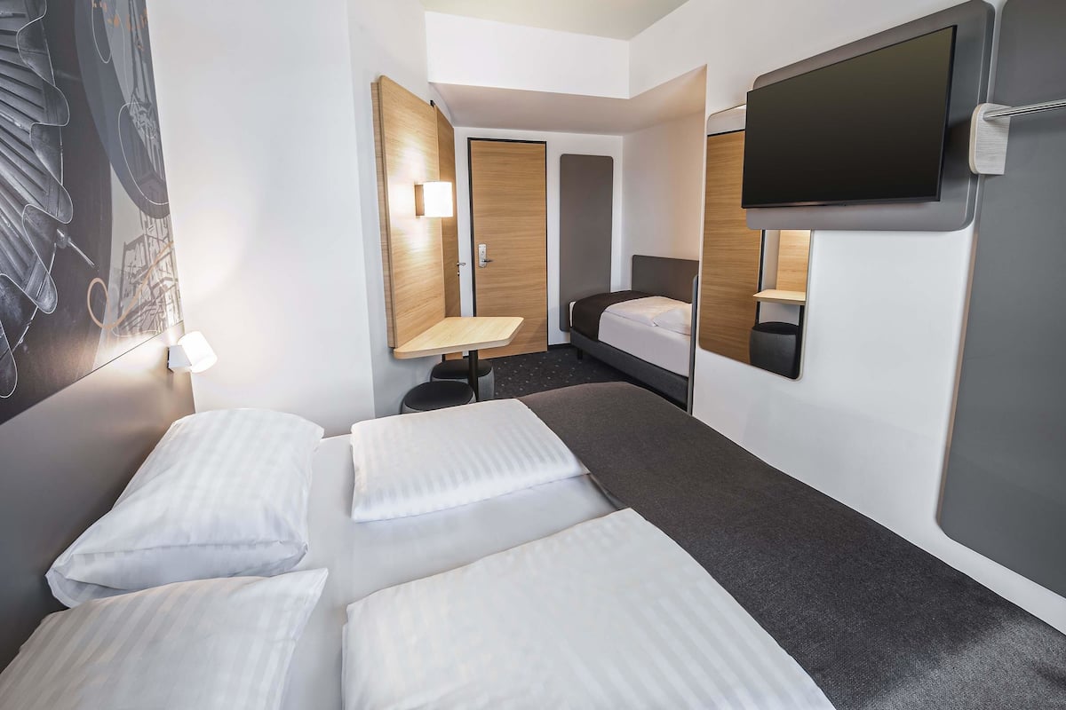 Habitación de hotel con cama, escritorio, silla, televisor y lámpara de pared.