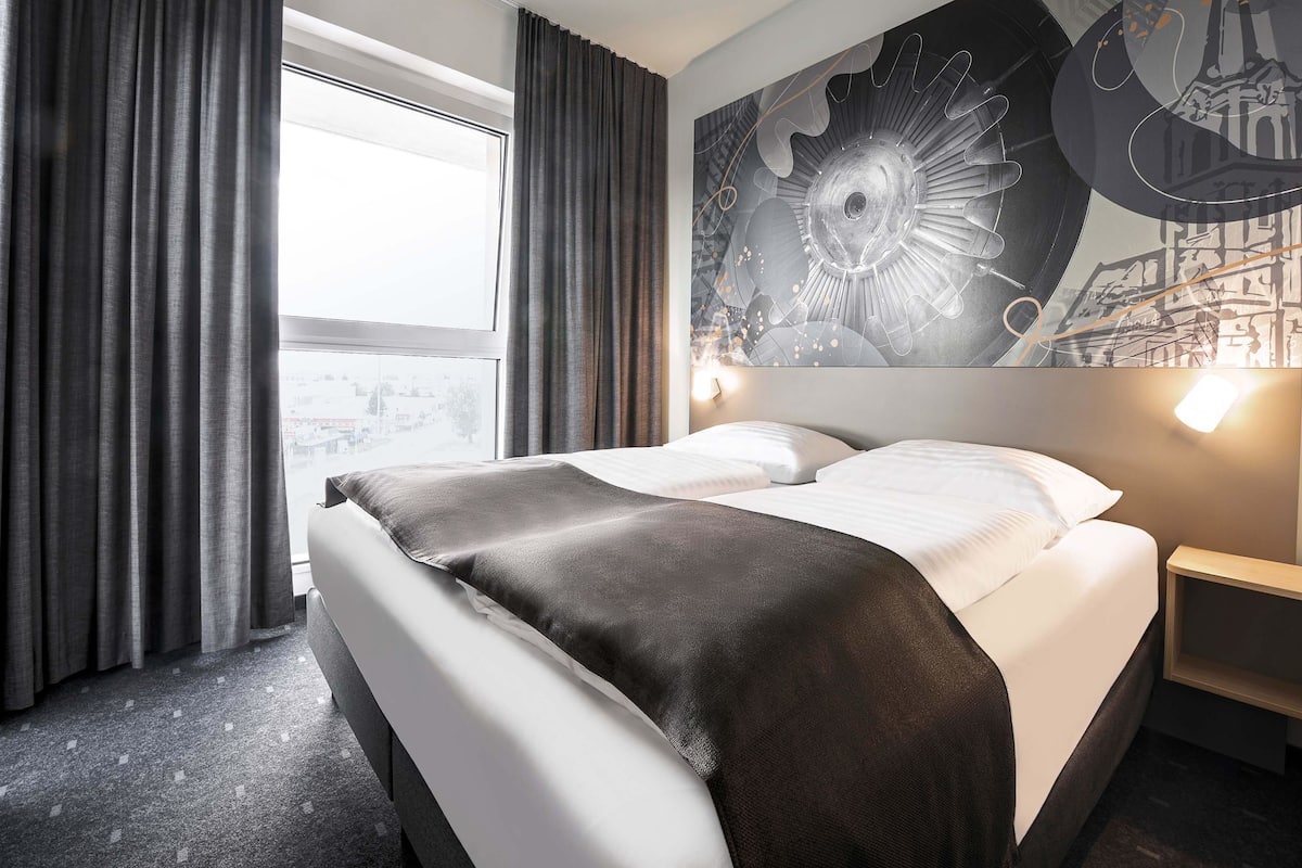 Habitación de hotel con una cama grande, vista a la ciudad por la ventana y un cuadro abstracto moderno en la pared.