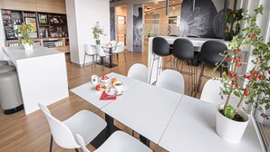 Un espace repas moderne avec des tables et des chaises blanches, un bar à café en arrière-plan et une plante en pot sur la table.
