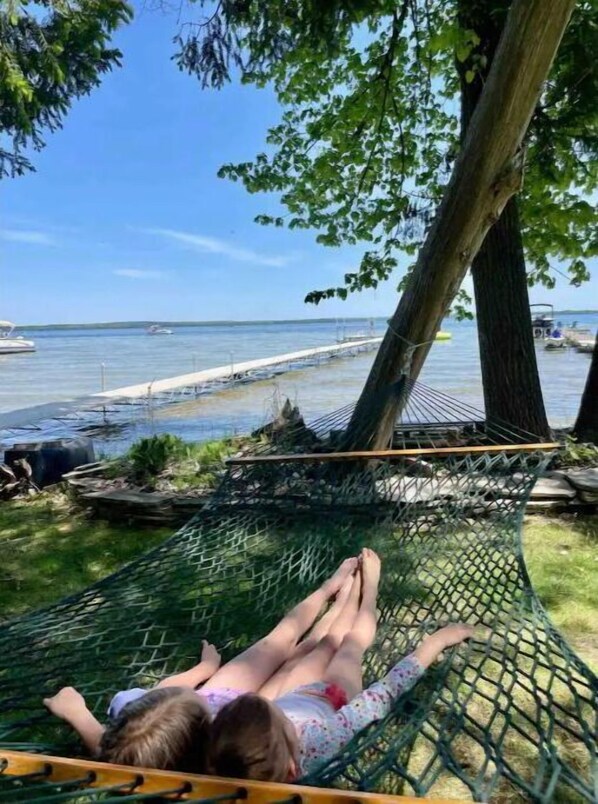 Relax in a hammock suspended between trees, overlooking a serene body of water with boats and a dock in the distance.