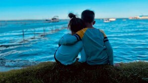 Un couple est assis sur une pelouse, d’où il a une vue sur une étendue d’eau où l’on aperçoit des bateaux au loin.